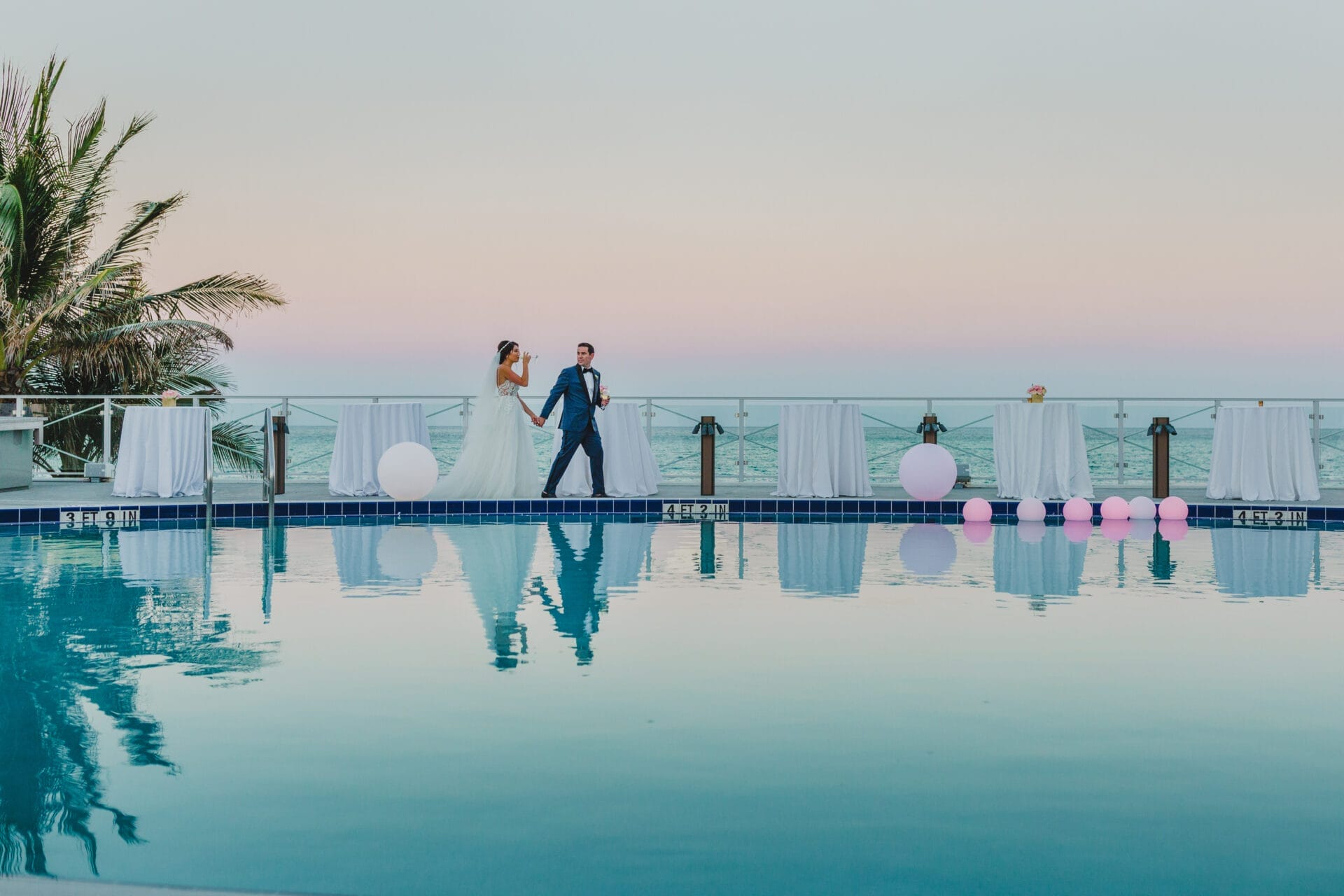 A bride and groom walk hand in hand along a poolside with the ocean in the background during sunset at Eden Roc, a luxurious Miami wedding venue. The serene water reflects the couple and the pastel sky, while elegant decor and soft lighting create a romantic atmosphere, making this an ideal location for a waterfront wedding in Miami.