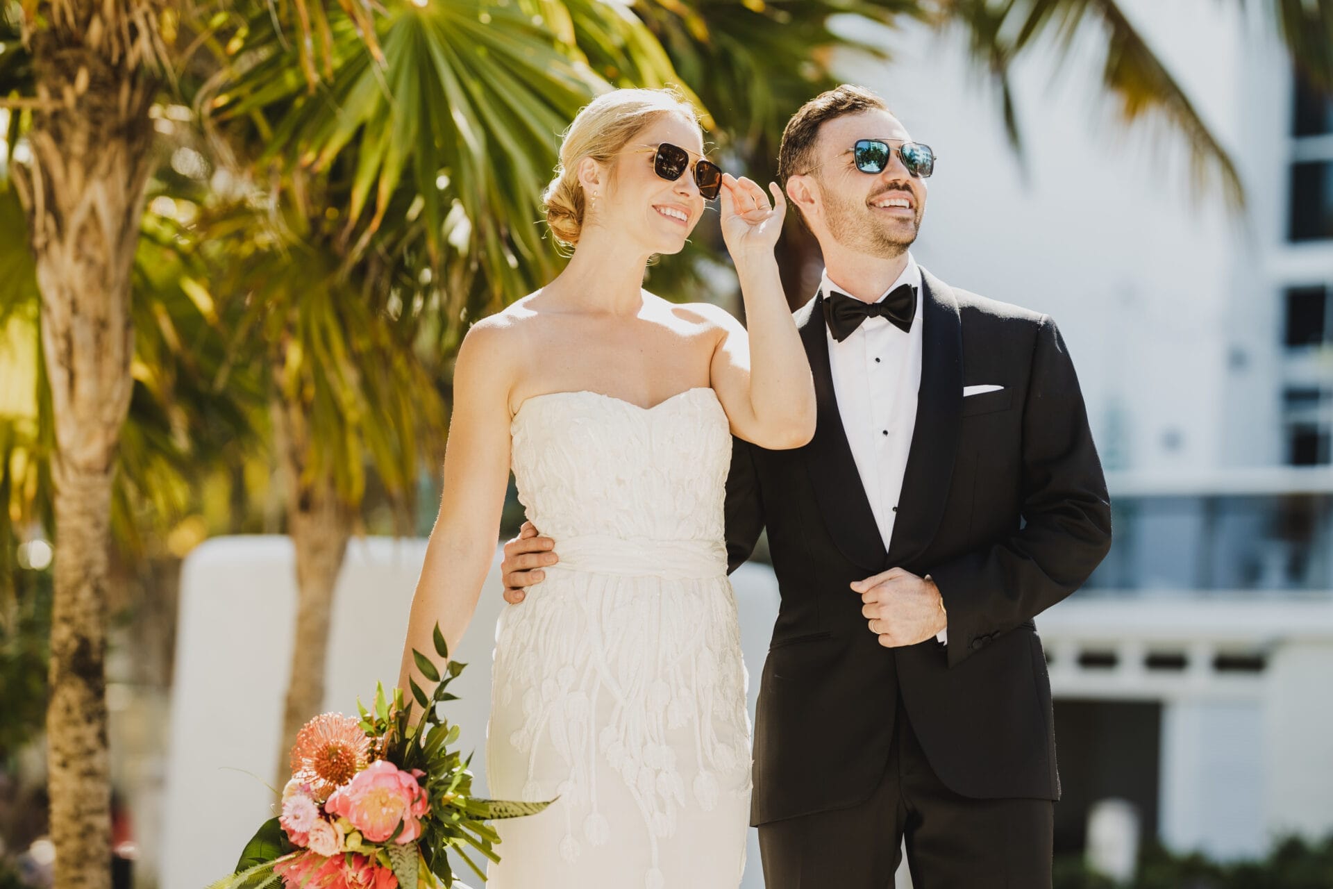 A bride and groom, both wearing stylish sunglasses, smile as they walk together in the sunshine, surrounded by palm trees at the Andaz Miami Beach. The bride carries a vibrant bouquet of pink and coral flowers, and the groom is dressed in a classic black tuxedo. This moment captures the relaxed and chic vibe of the Andaz, making it a perfect Miami wedding venue for couples looking for a modern and tropical setting.