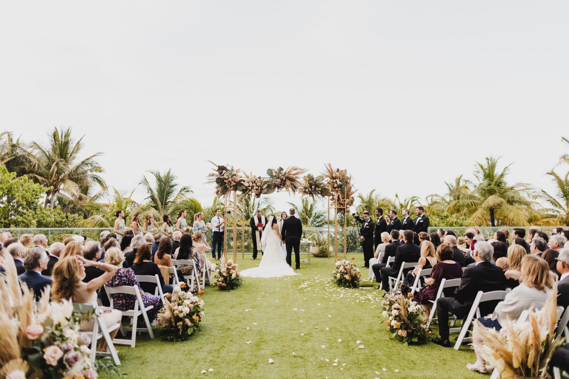 A bride and groom stand at the altar surrounded by lush greenery and tropical palm trees during an outdoor ceremony at 1 Hotel South Beach, a premier Miami wedding venue. The aisle is lined with floral arrangements, and guests are seated on either side, creating an intimate and elegant setting for this beachfront wedding in Miami.