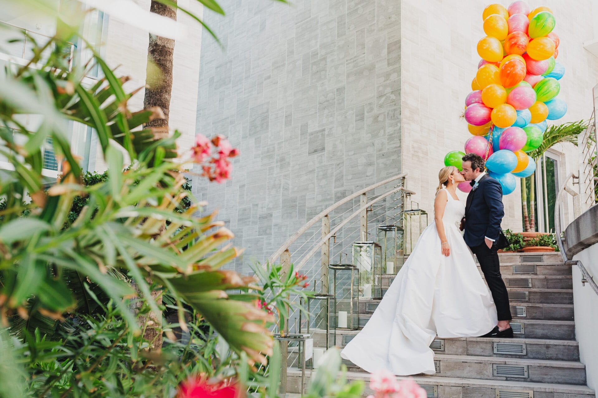 A bride and groom share a kiss on a staircase surrounded by lush greenery and colorful balloons at The Miami Beach EDITION, one of the top Miami wedding venues known for its modern elegance and luxurious setting.