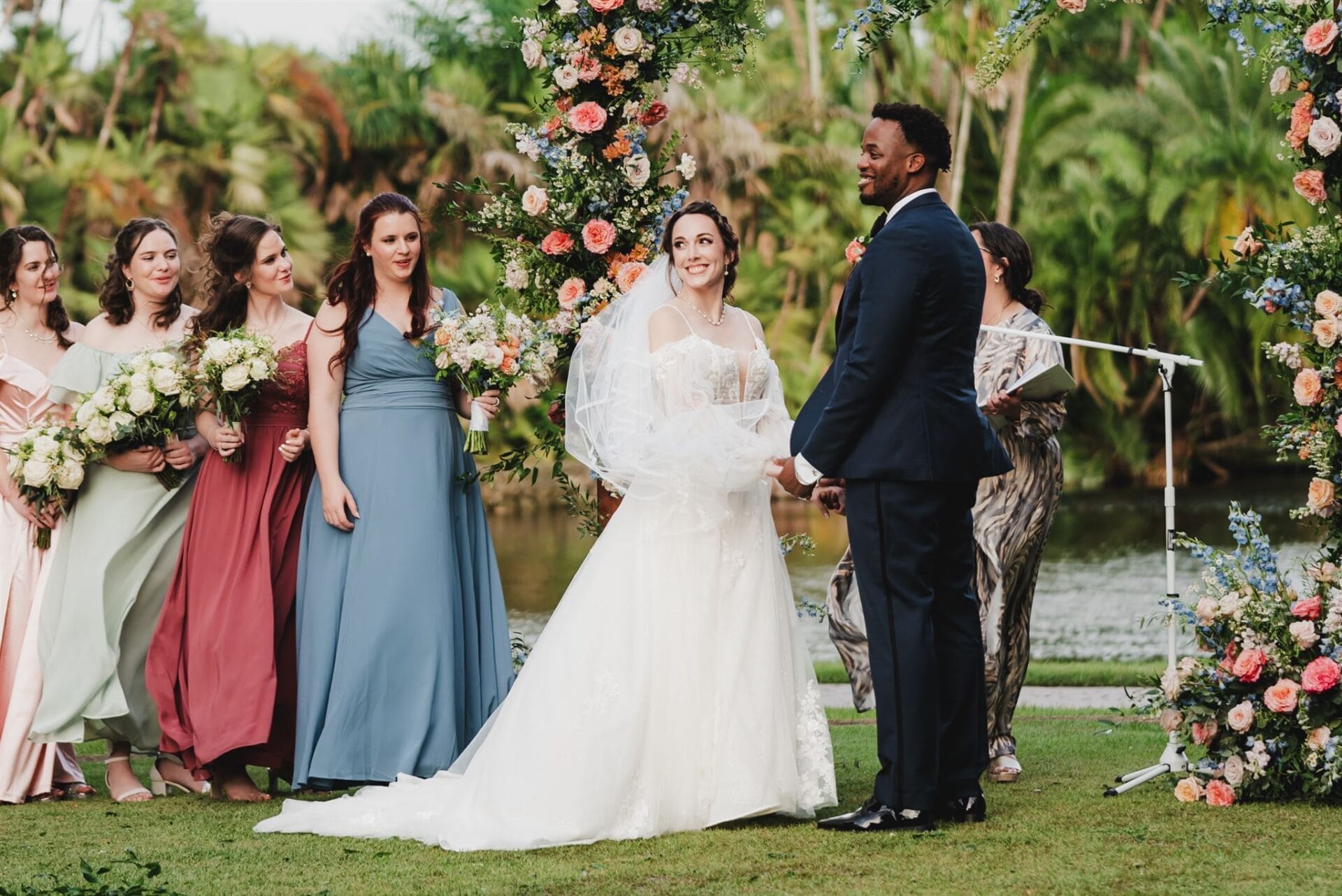 Bride and groom exchanging vows during an outdoor wedding ceremony, surrounded by bridesmaids in colorful dresses and a floral arch.