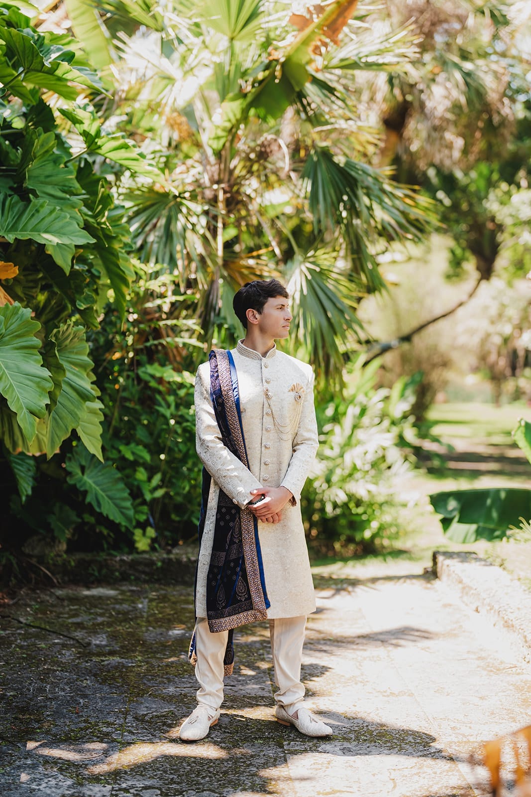 Man in traditional Indian attire standing in a lush garden, wearing a cream-colored sherwani with intricate designs and a dark blue dupatta draped over his shoulder. The background features vibrant green tropical plants, creating a serene outdoor setting.