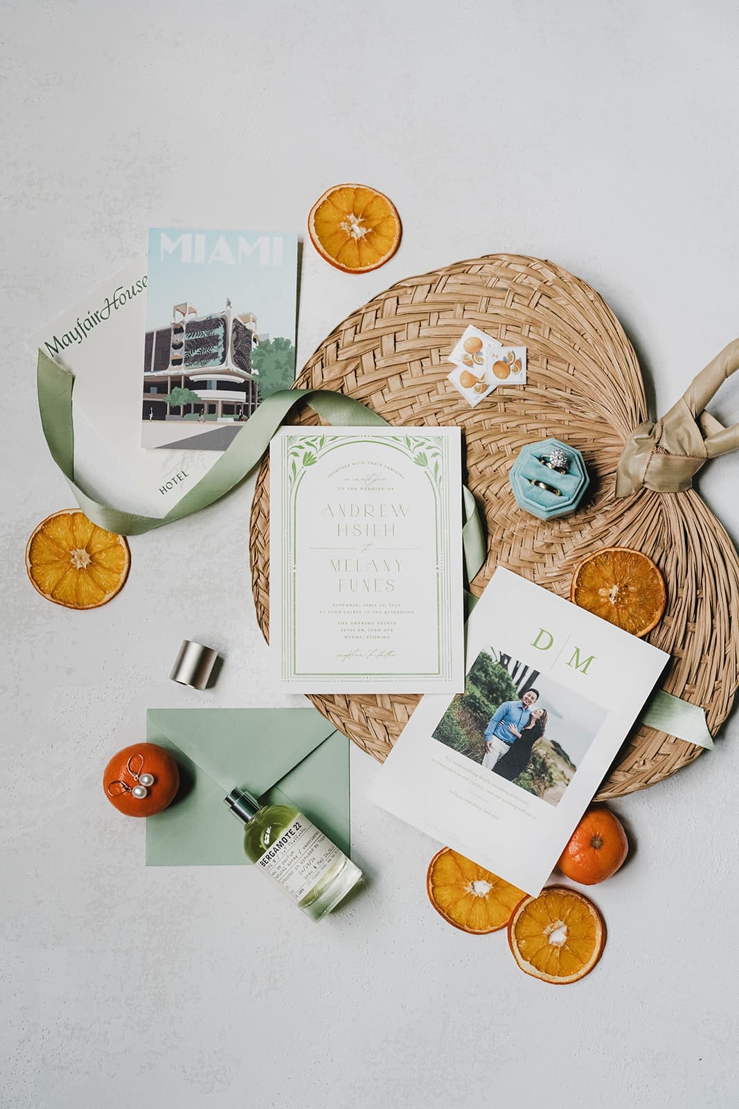 A flat lay image featuring wedding invitations, a green ribbon, a bottle of fragrance, and decorative oranges on a woven placemat. The layout includes a Miami hotel brochure, personalized invitations for Andrew Hsieh and Melany Funes, and an engagement photo card. The arrangement highlights a stylish and festive wedding planning theme.