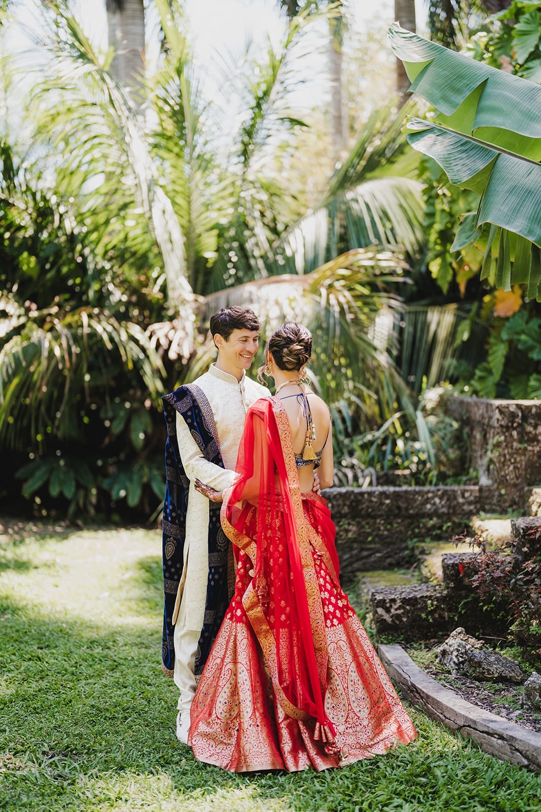 Couple dressed in traditional Indian attire, standing in a lush garden. The woman wears a red and gold lehenga with a matching dupatta, while the man is in a cream-colored kurta with a blue and gold shawl. They appear joyful and engaged in a moment together, surrounded by tropical plants.