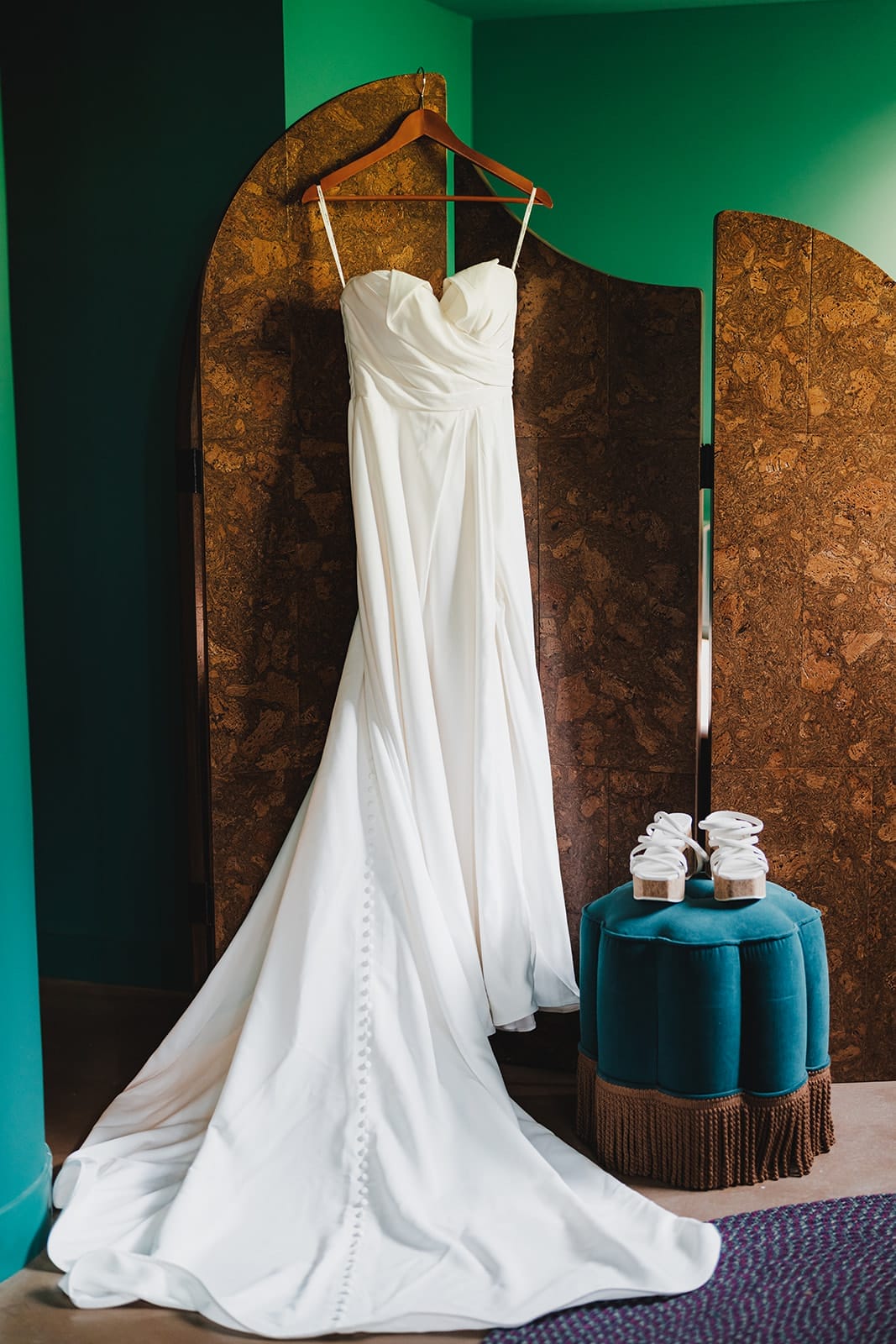 Elegant white wedding dress hanging on a wooden hanger against a cork backdrop, with a pair of stylish white platform shoes displayed on a teal ottoman nearby.