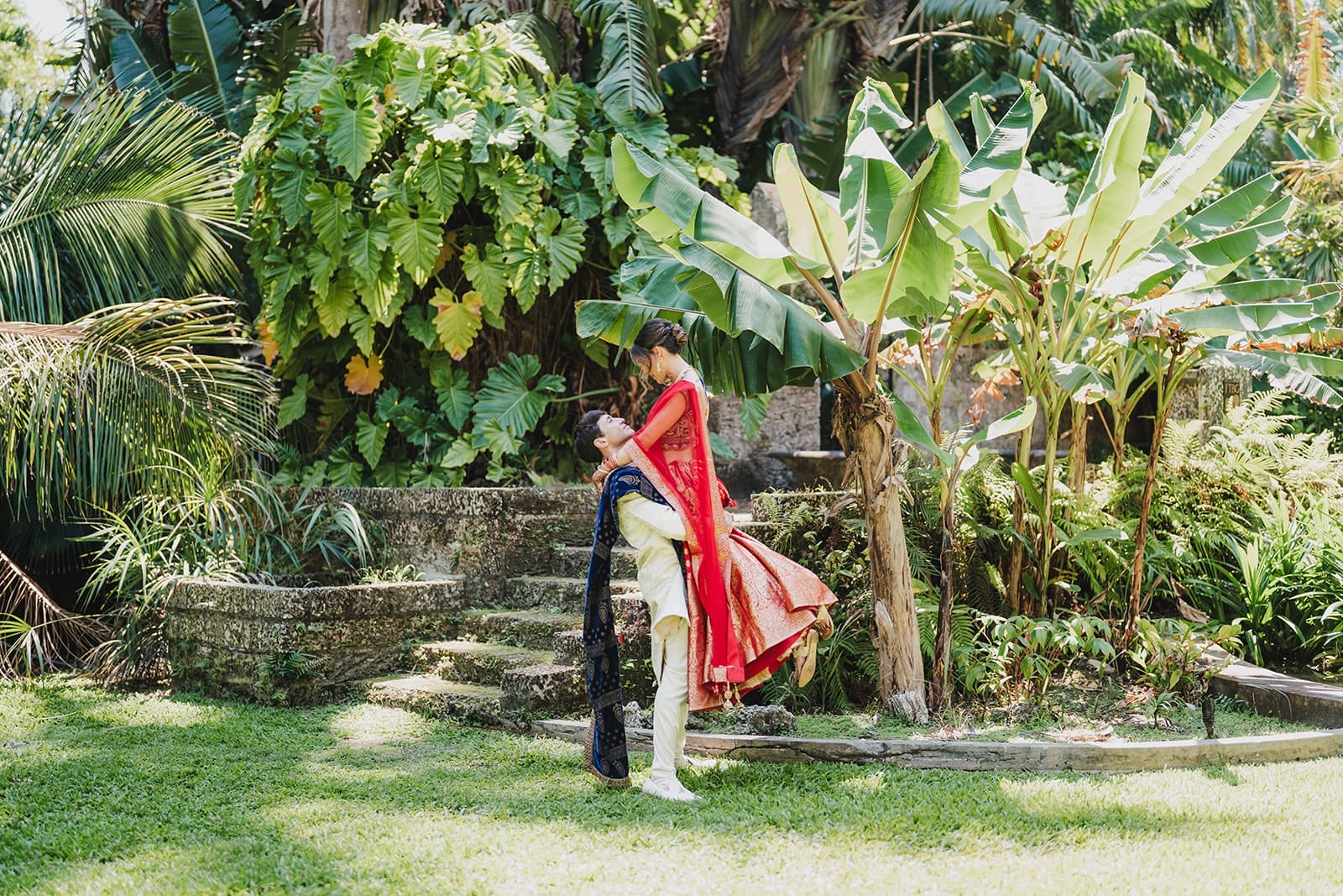 Couple in traditional Indian attire poses in a lush garden, with the man lifting the woman joyfully against a backdrop of tropical plants and stone steps.
