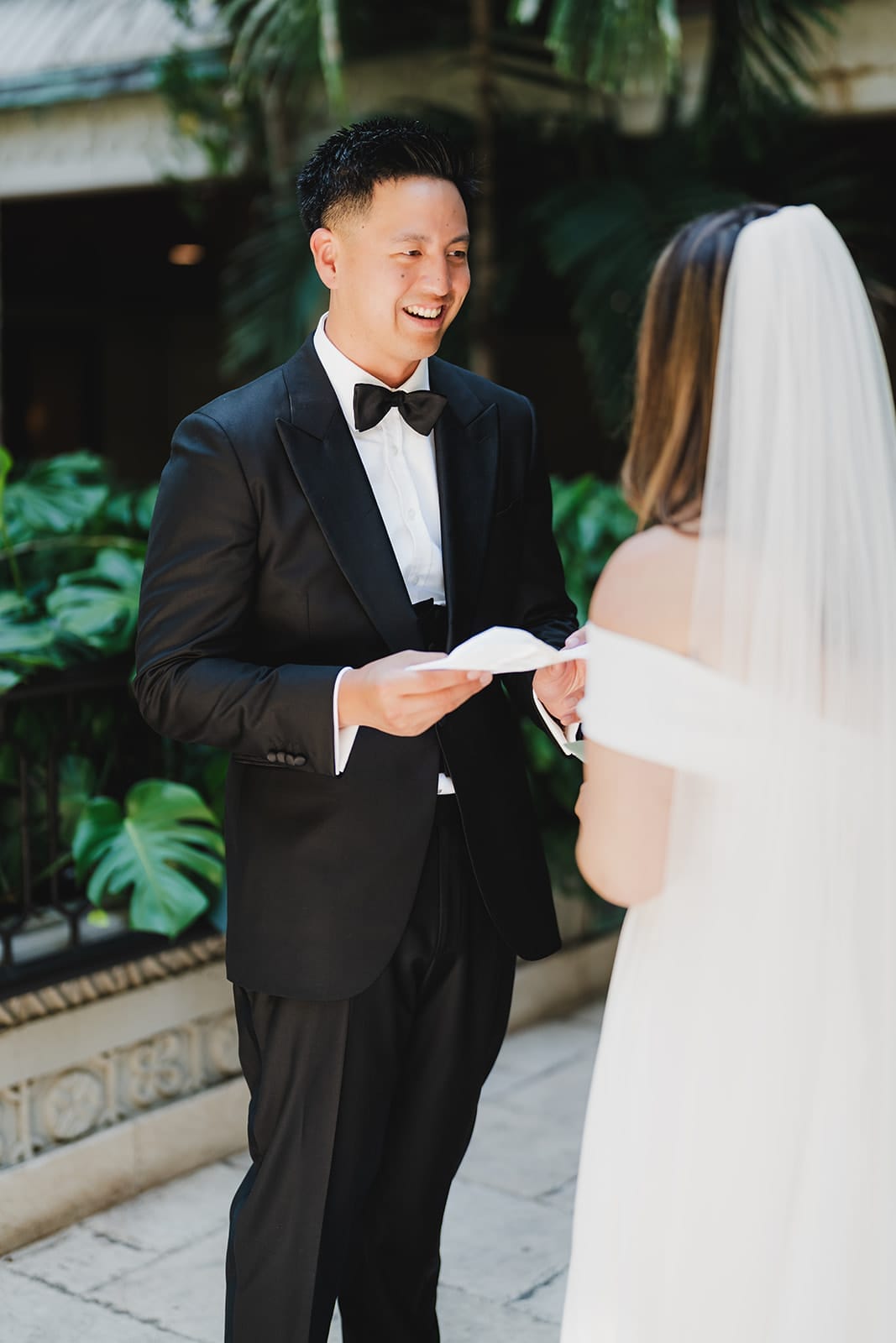 Groom in a black tuxedo smiles while reading vows to his bride in a wedding setting surrounded by lush greenery.