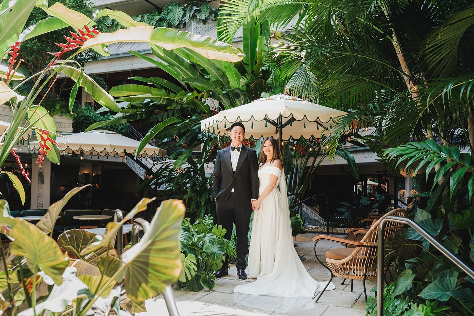 Bride and groom standing together in a lush, tropical garden setting, surrounded by large green plants and decorative umbrellas, showcasing a romantic wedding atmosphere. The bride wears a white off-the-shoulder gown, while the groom is dressed in a black tuxedo.