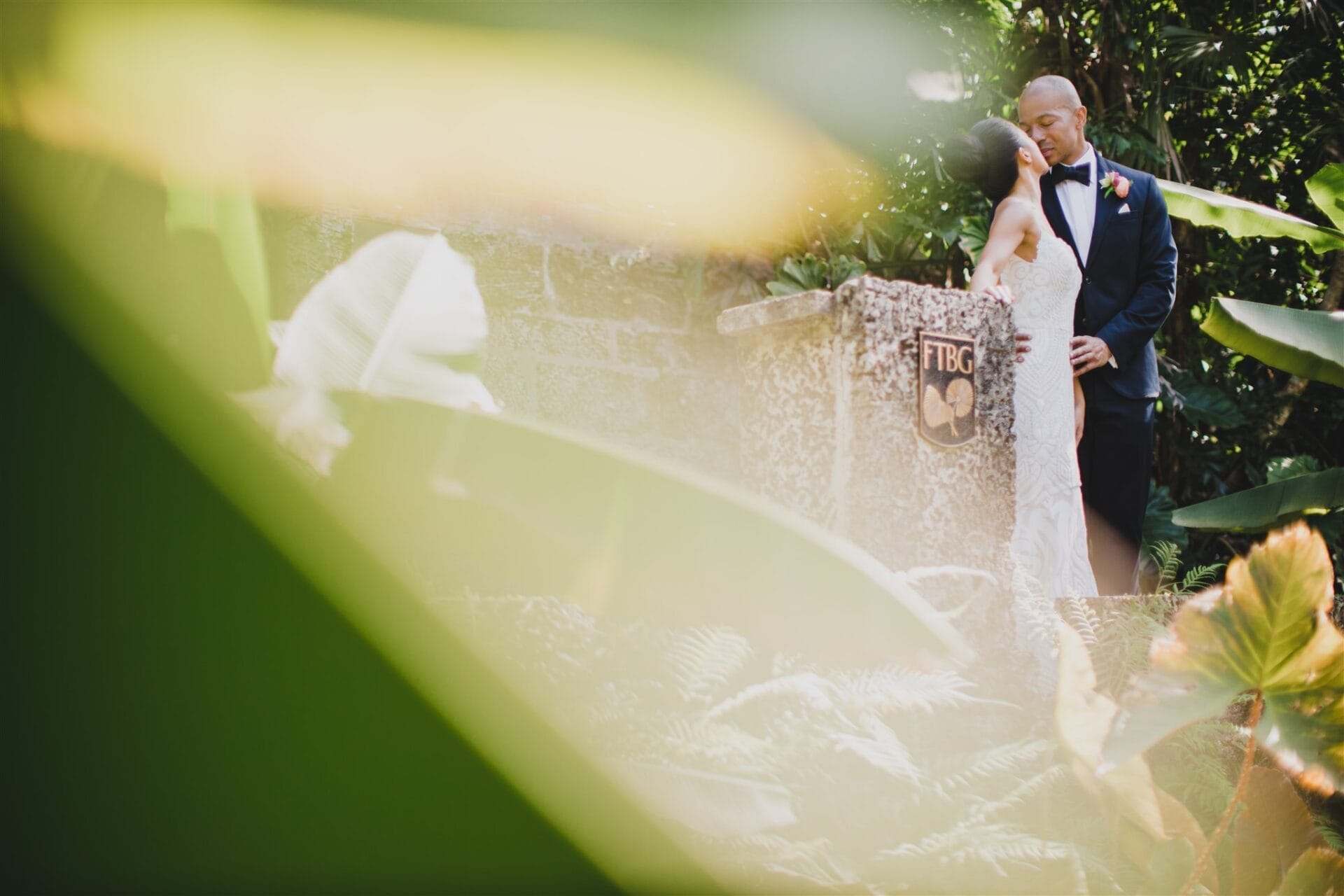 Couple in formal attire sharing a romantic moment at an outdoor wedding ceremony, surrounded by lush greenery and tropical plants. The scene features a decorative stone altar with an emblem, capturing the essence of a beautiful wedding setting.