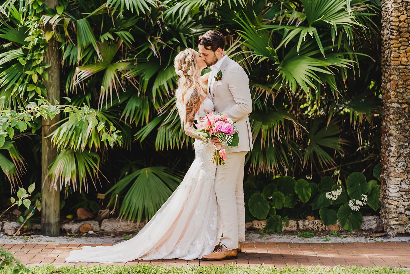 Bride and groom sharing a kiss in a tropical garden setting, surrounded by lush green palm leaves. The bride wears a fitted lace wedding dress and holds a vibrant bouquet, while the groom is dressed in a light-colored suit.