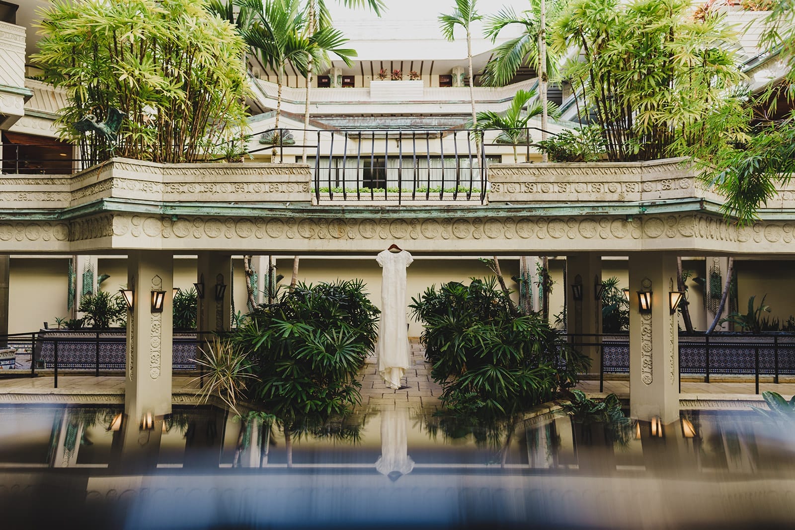 Interior view of a tropical hotel atrium featuring lush greenery, decorative architecture, and a serene reflective pool, with a white dress displayed prominently among the plants.