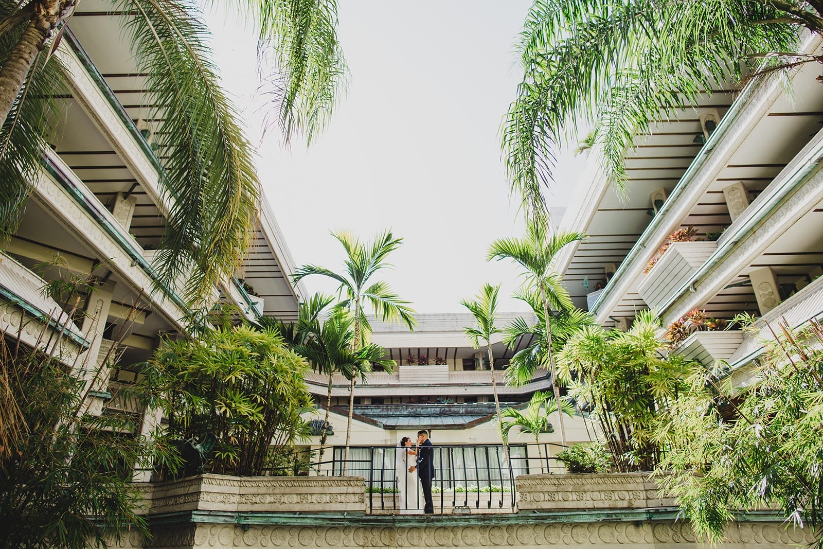 Couple embracing in a lush courtyard surrounded by tropical plants and modern architecture, capturing a romantic moment in a serene outdoor setting.