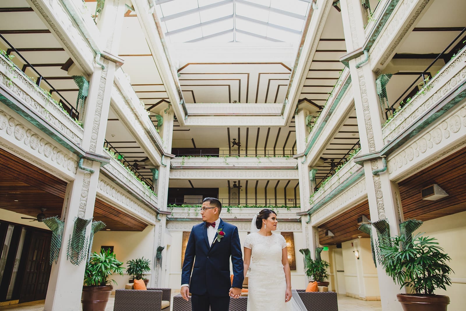 Bride and groom standing hand in hand in a beautifully designed indoor venue, featuring intricate architectural details and greenery. The couple is dressed elegantly, with the bride in a lace wedding gown and the groom in a classic tuxedo, capturing a moment of intimacy in a romantic setting.