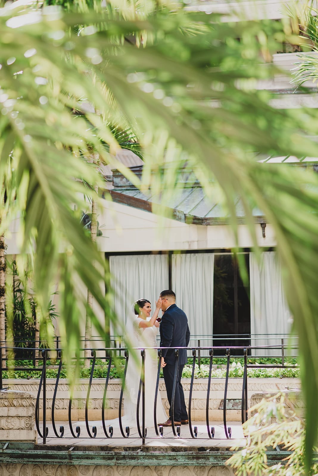Bride and groom share a tender moment on a balcony surrounded by lush greenery, capturing the essence of romance and celebration in a wedding setting.