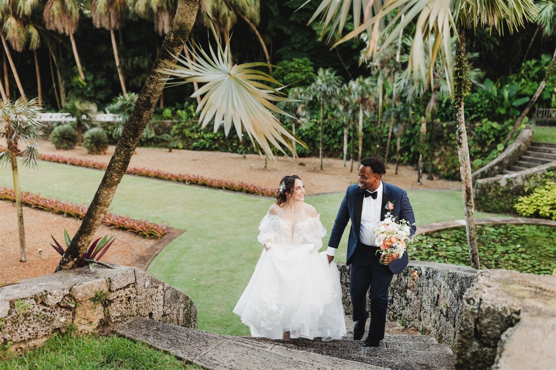 Bride and groom walking hand in hand down stone steps in a lush garden setting, surrounded by tropical plants and colorful flower beds. The bride is wearing a white wedding gown, and the groom is dressed in a navy tuxedo, holding a floral bouquet.