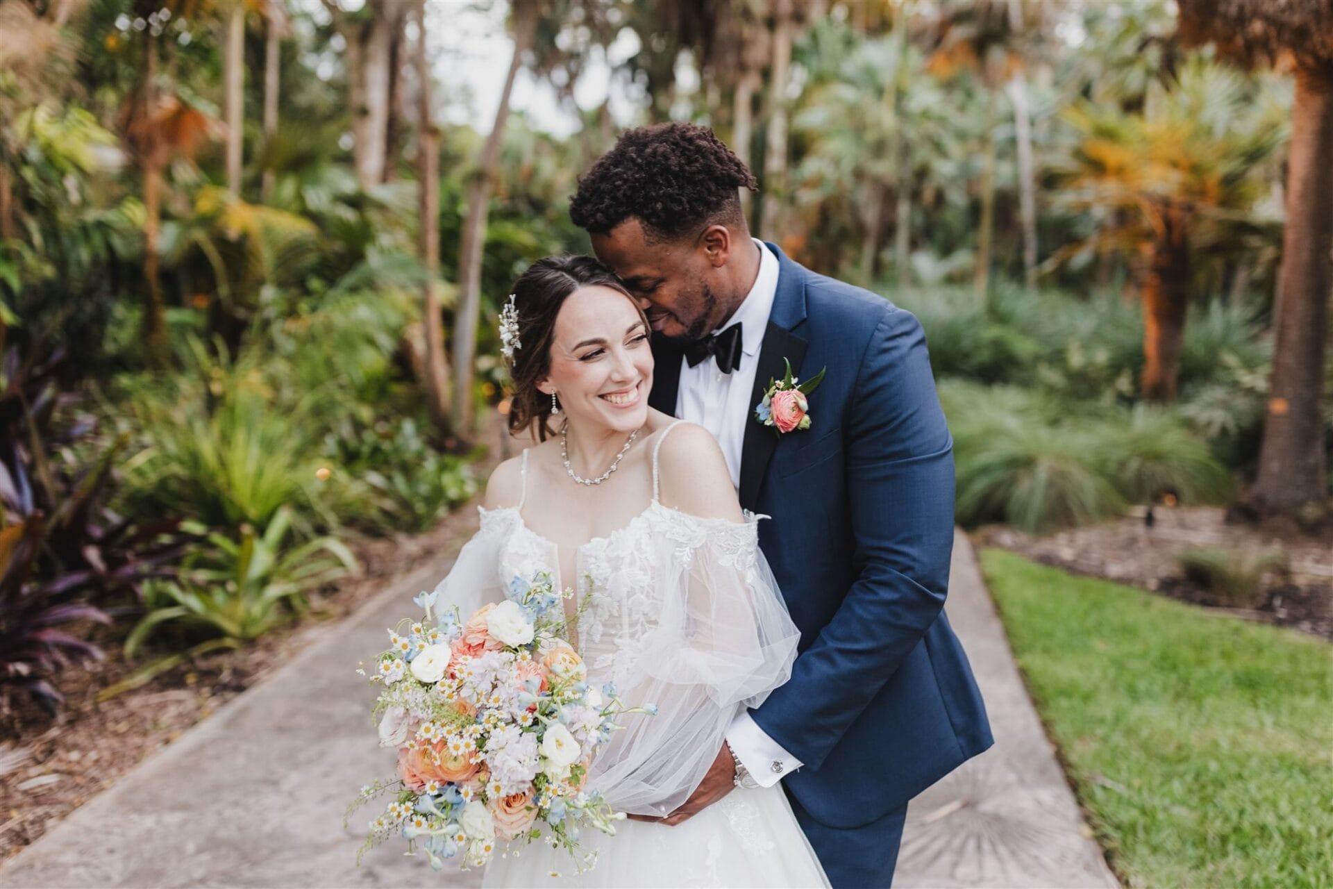 A joyful bride and groom embrace in a lush garden setting, with the bride holding a colorful bouquet of flowers. The groom, in a navy suit with a black bow tie, leans in close to the bride, who wears an elegant wedding dress with sheer sleeves and a floral hair accessory. The background features vibrant greenery, creating a romantic atmosphere for their outdoor wedding.