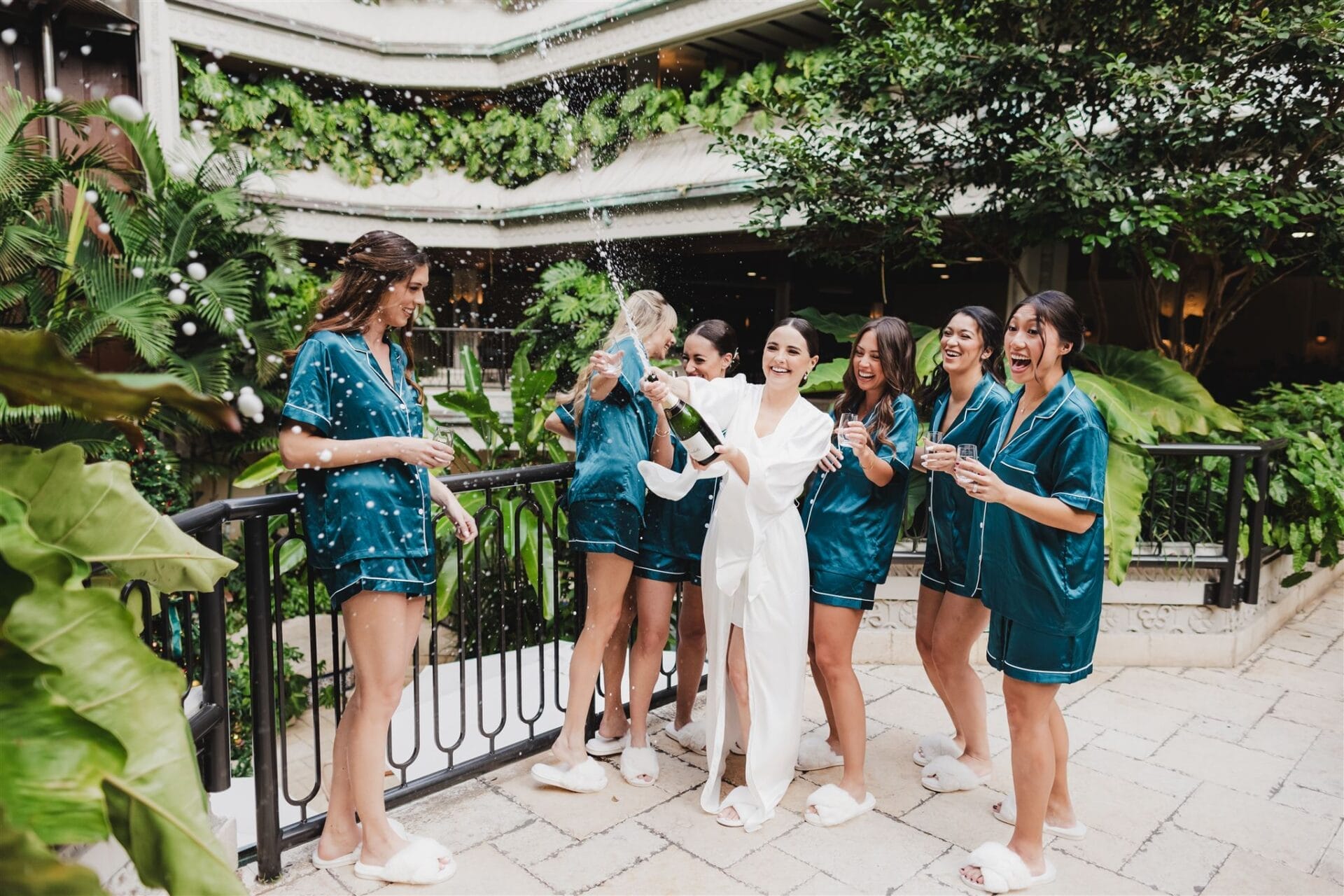 Group of women in matching teal pajamas celebrating with champagne outdoors, surrounded by lush greenery. One woman in a white robe is joyfully opening a champagne bottle while the others cheer and hold glasses.