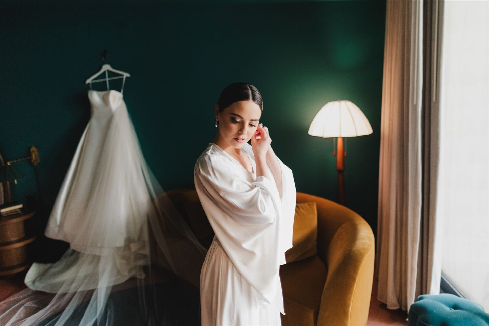 Bride getting ready in a stylish room, wearing a white robe while adjusting her earrings, with a wedding dress hanging in the background. The setting features a green wall, a modern lamp, and a cozy yellow sofa, creating an elegant atmosphere for wedding preparations.