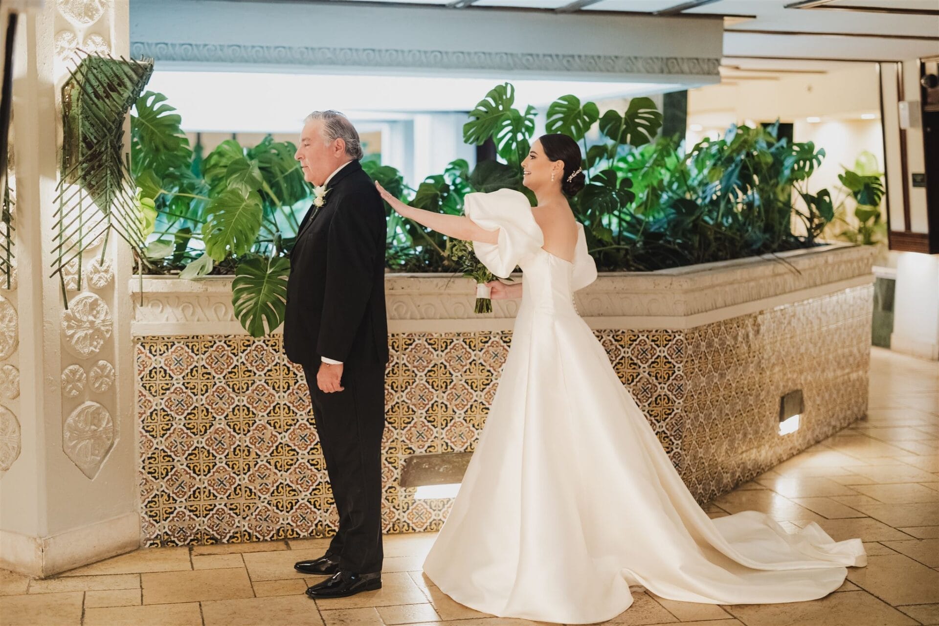 Bride in a white wedding dress surprises her father in a formal black suit, set against a backdrop of lush greenery and decorative tilework in an indoor venue.