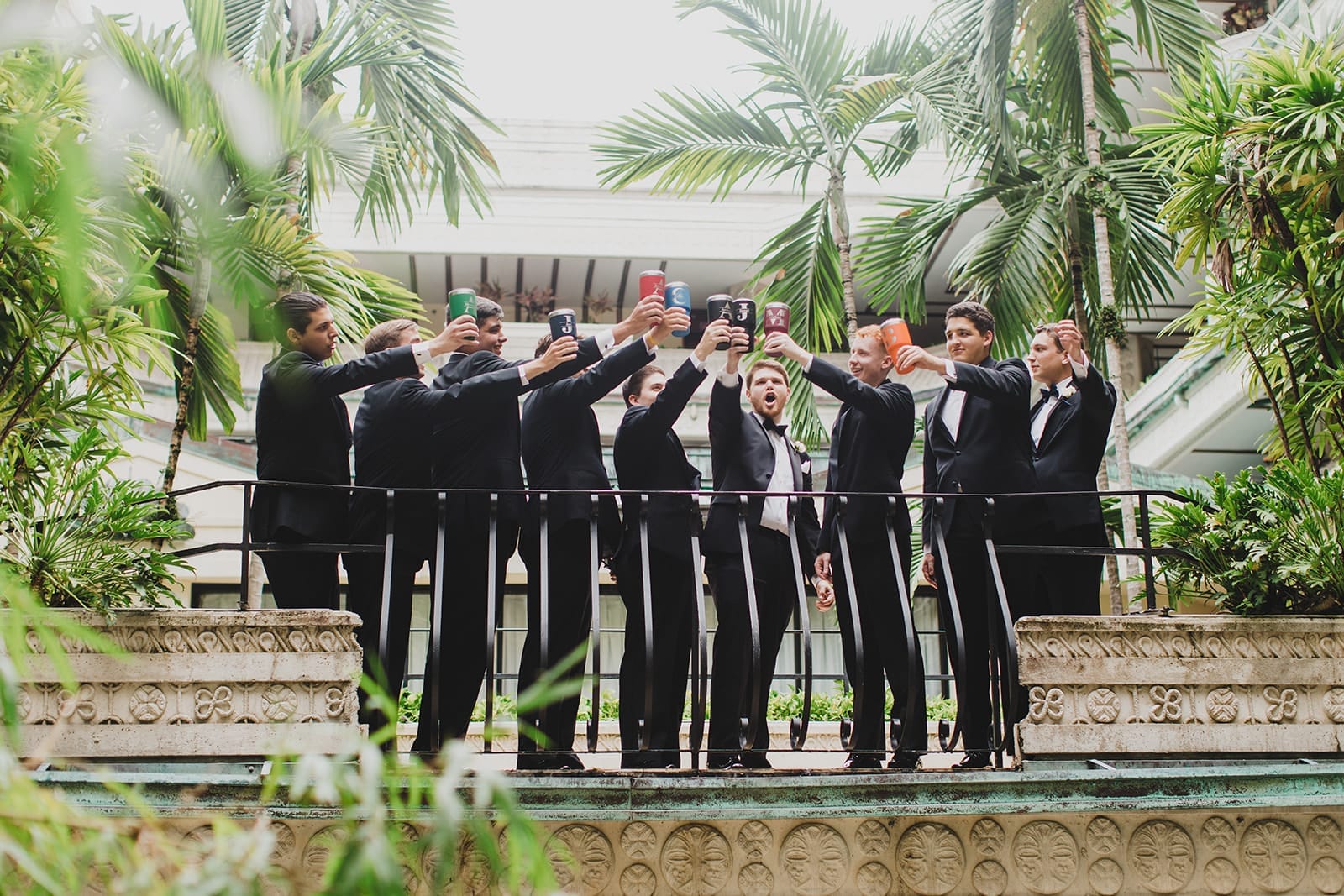 Groom and groomsmen in tuxedos celebrating on a balcony, raising drinks in a lush garden setting. The group showcases camaraderie and joy, perfect for wedding or event photography.