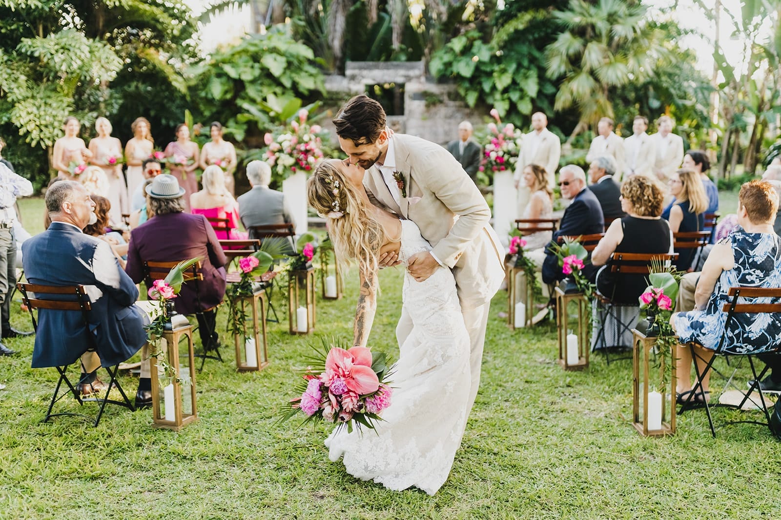 Bride and groom share a romantic kiss during their outdoor wedding ceremony, surrounded by guests seated on wooden chairs adorned with floral arrangements. The lush greenery and vibrant floral decorations enhance the intimate atmosphere of the event.