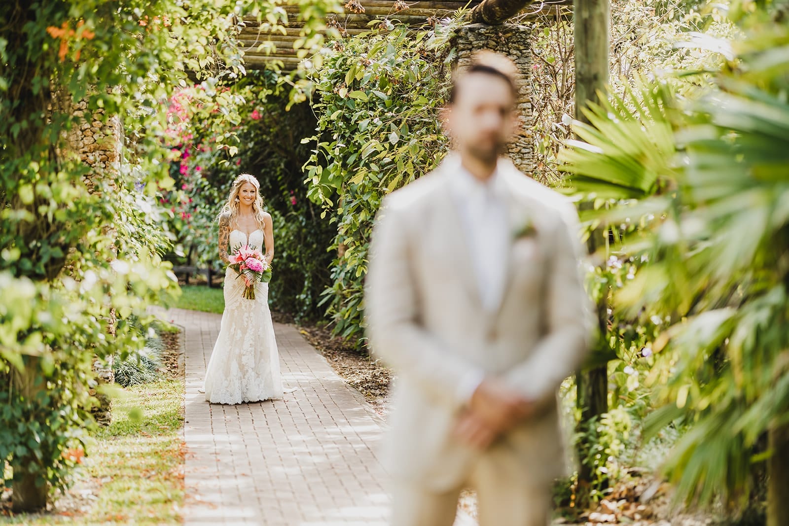 Bride in a lace wedding dress holding a bouquet of pink flowers, walking down a garden path towards a groom in a beige suit. Lush greenery and vibrant flowers frame the scene, creating a romantic atmosphere.