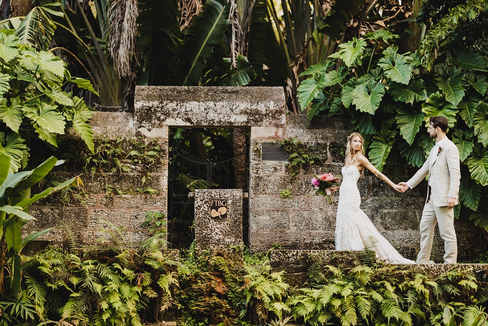 Bride and groom holding hands in a lush tropical garden, standing in front of an old stone structure adorned with greenery. The bride wears a white lace wedding gown and holds a bouquet of vibrant flowers, while the groom is dressed in a light-colored suit.