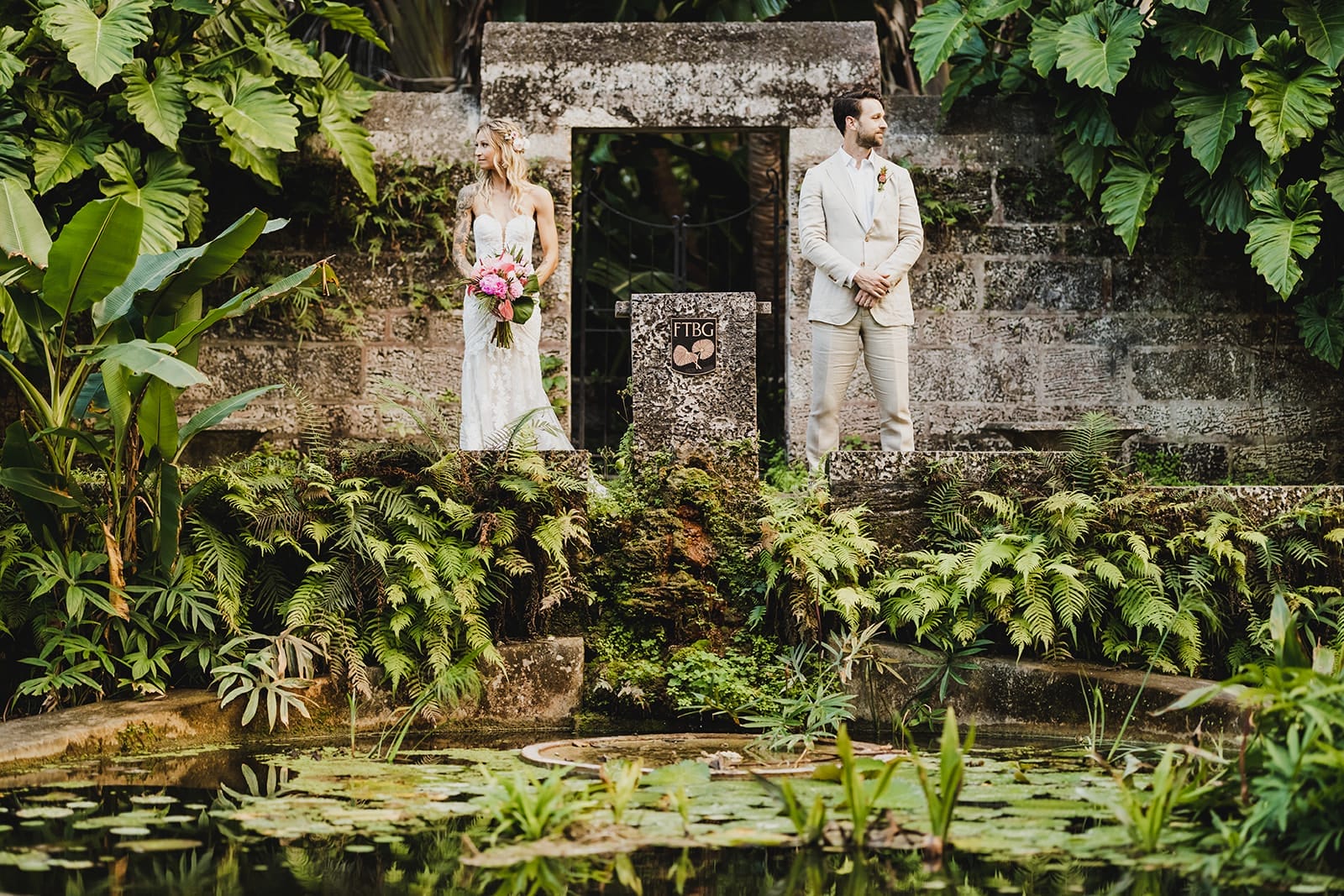 Bride and groom standing in a lush garden setting, with the bride in a white lace wedding dress holding a bouquet of pink flowers, and the groom in a light-colored suit. The scene features tropical plants and a pond with lily pads, creating a romantic backdrop for a wedding ceremony.