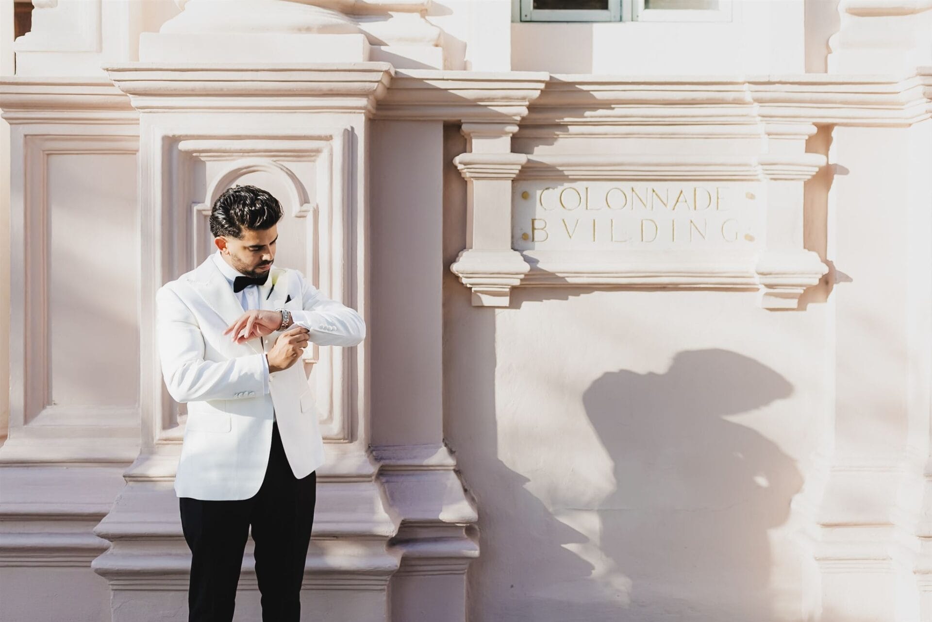 Man in a white tuxedo adjusting his watch in front of a pink wall with the text "Colonnade Building." The setting suggests a formal event or wedding.