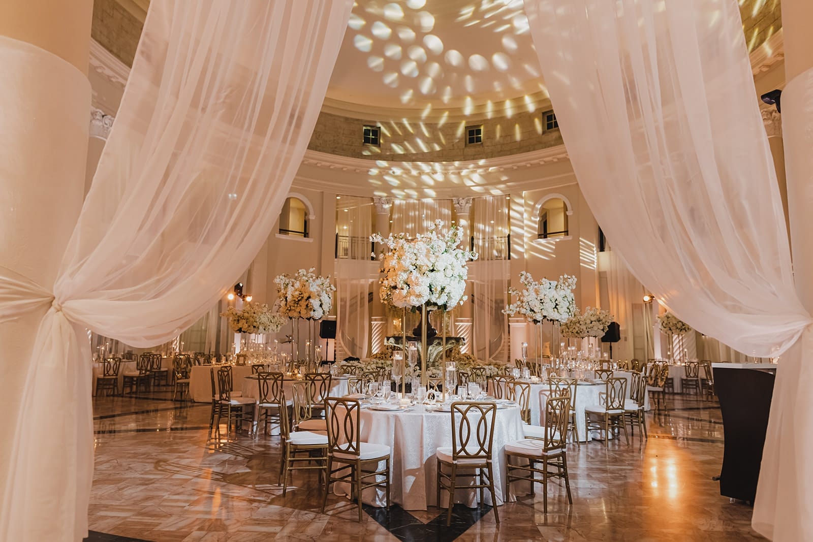 Elegant event space featuring round tables adorned with floral centerpieces, surrounded by gold chairs. Sheer drapes frame the room, highlighting the sophisticated ambiance created by soft lighting and decorative wall patterns. Ideal for weddings and formal gatherings.